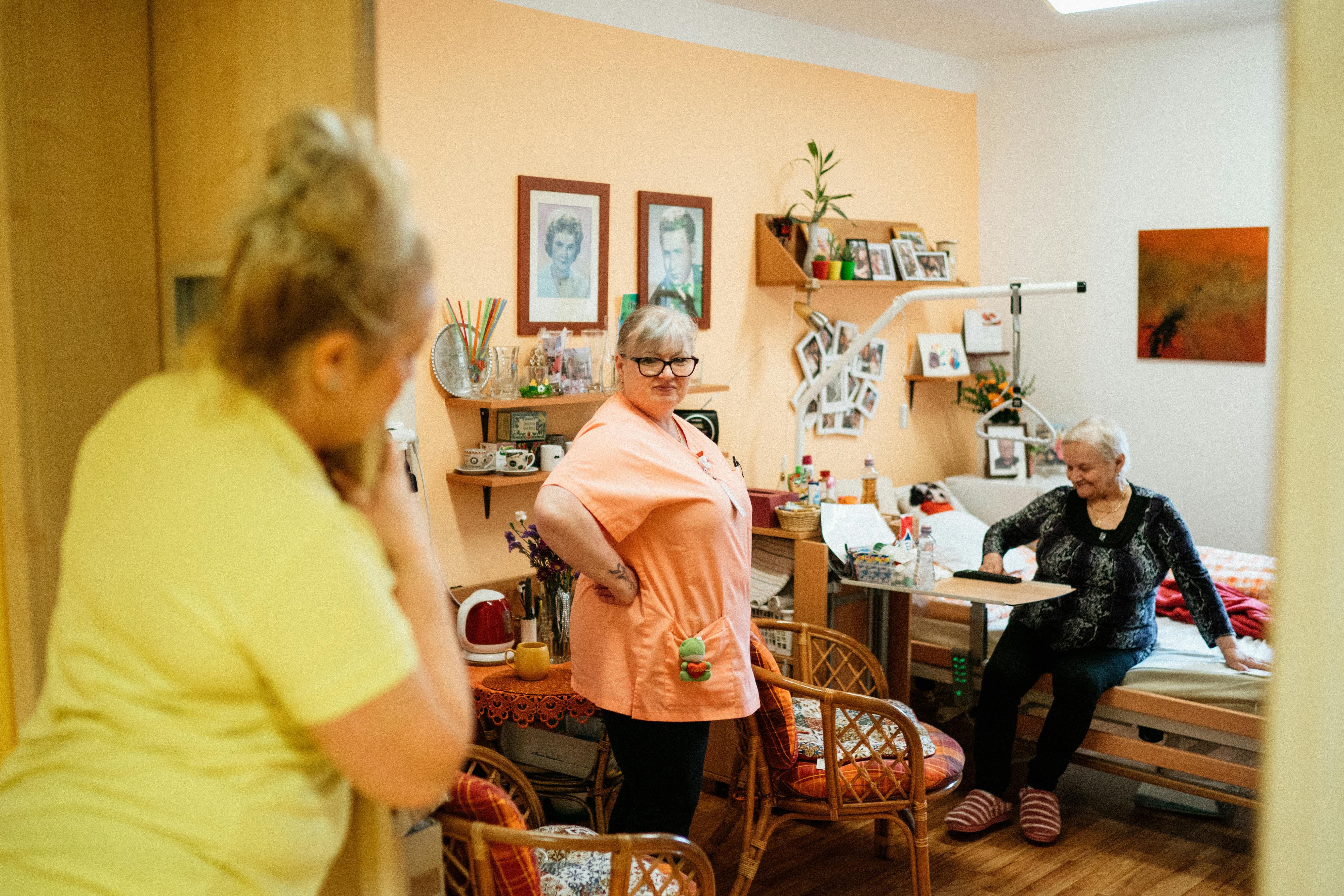 In the foreground, a woman with blonde hair, wearing a yellow top, is leaning against the doorframe. In the middle, a woman in an orange uniform with glasses stands smiling. Behind her, an elderly woman with short white hair sits on a bed, wearing a black patterned jumper.