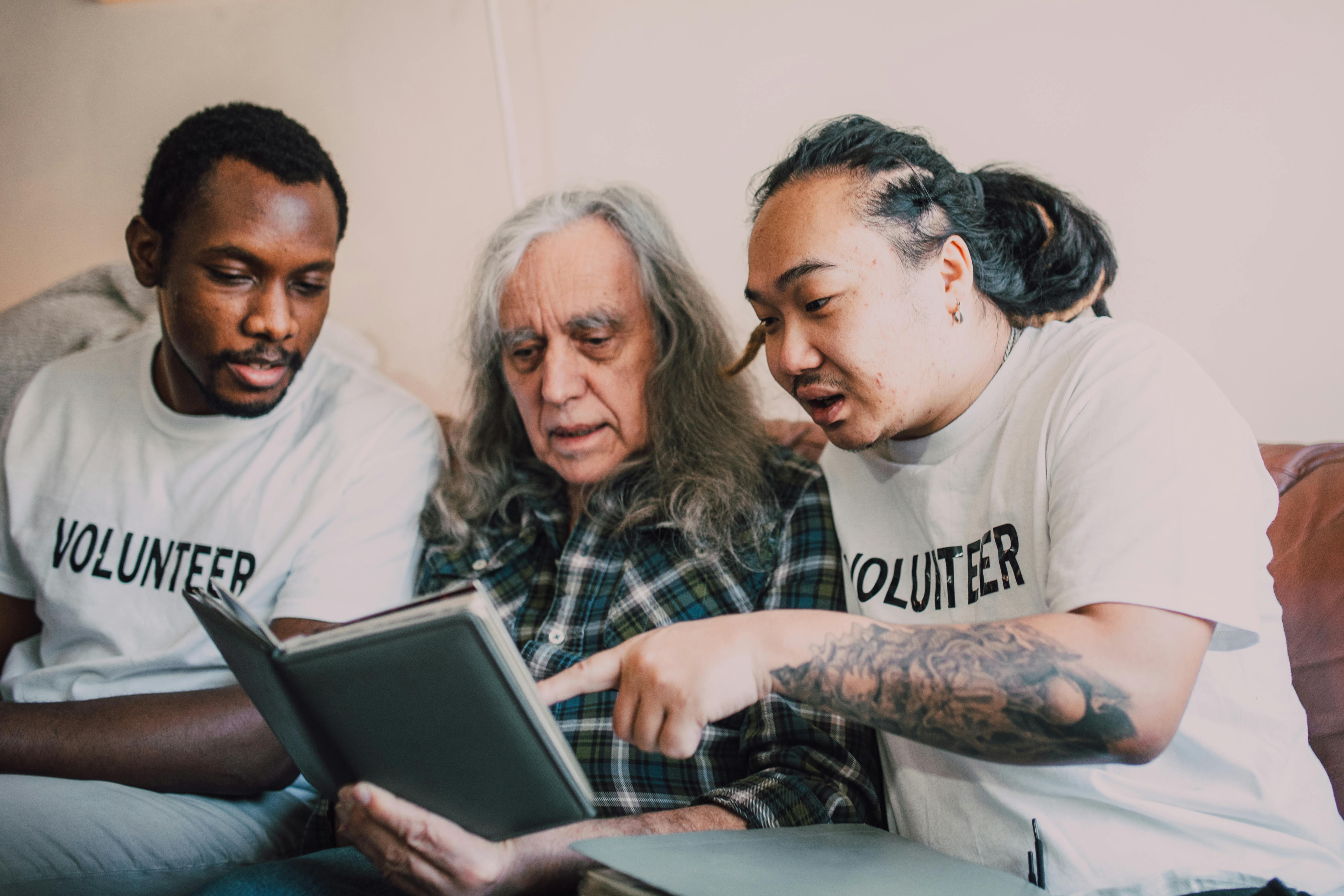 Two young volunteers sitting on a couch with an elderly man, all looking at a photo album or book. The volunteers wear white shirts labeled 'VOLUNTEER' and appear engaged and supportive as they help the elderly man.