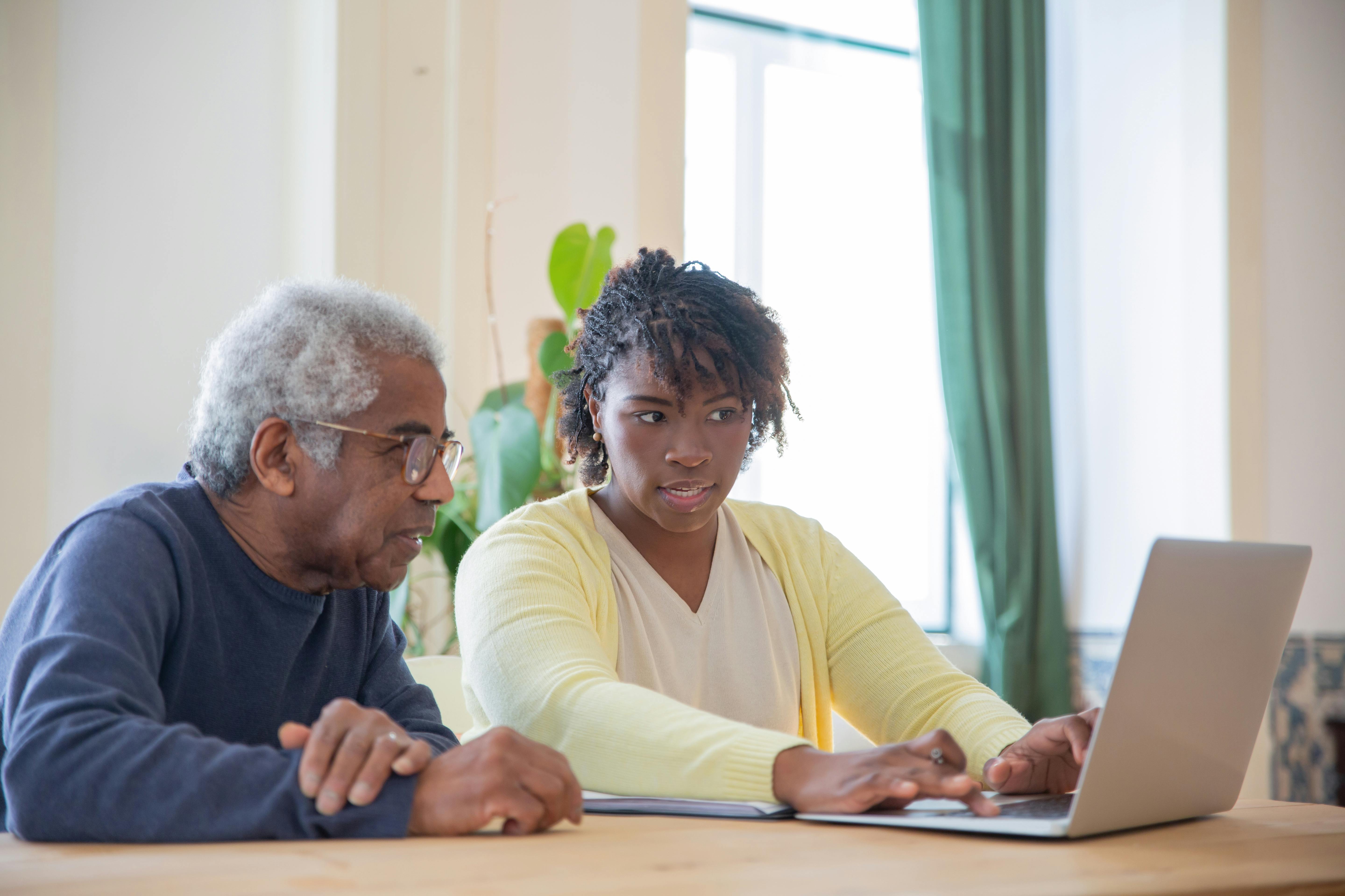 A young woman helps an elderly man use a laptop at a table.