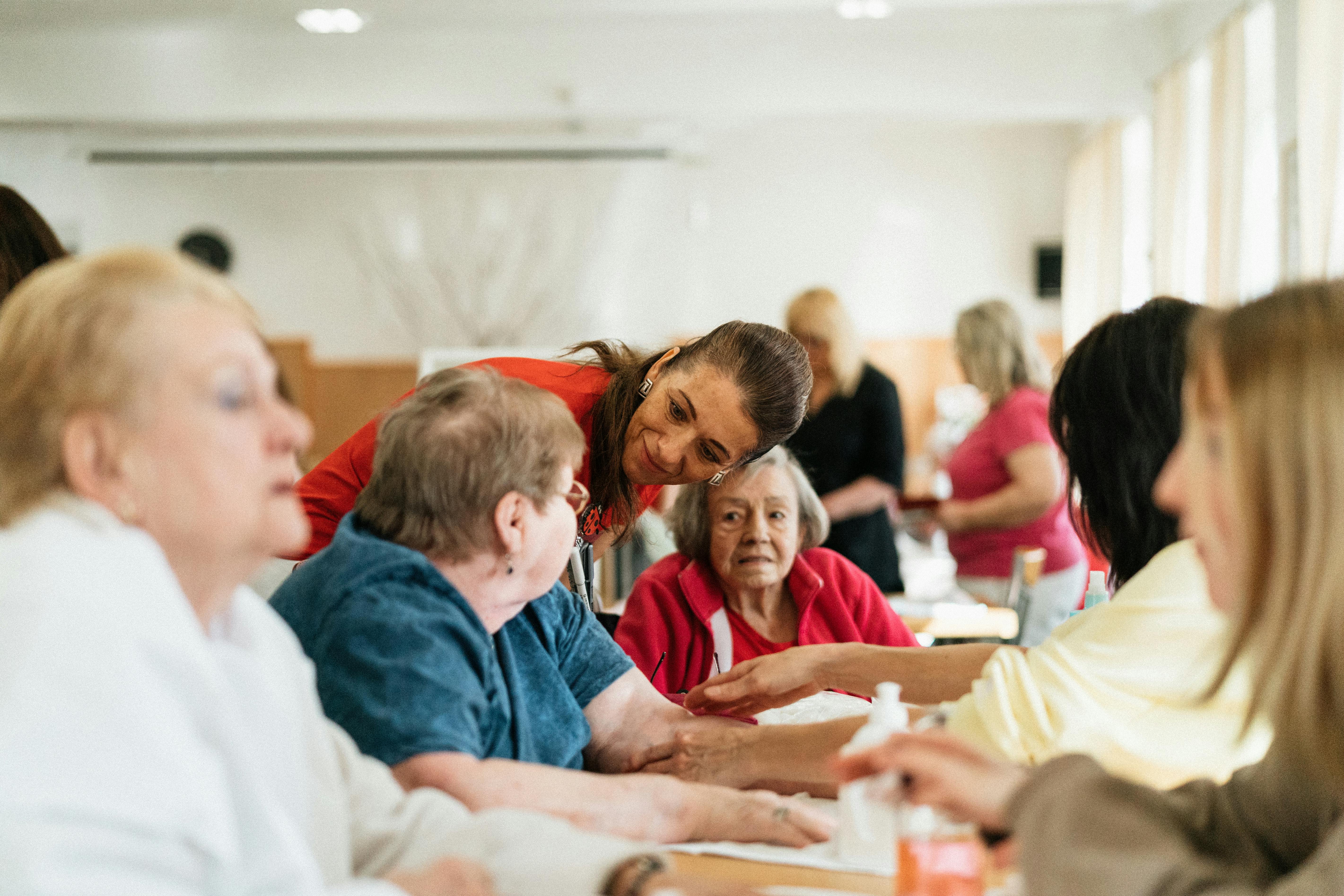 A caregiver in a red shirt warmly interacting with a group of elderly women seated at a table in a community or nursing home setting. The atmosphere is friendly and supportive, with people engaged in conversation and activities.