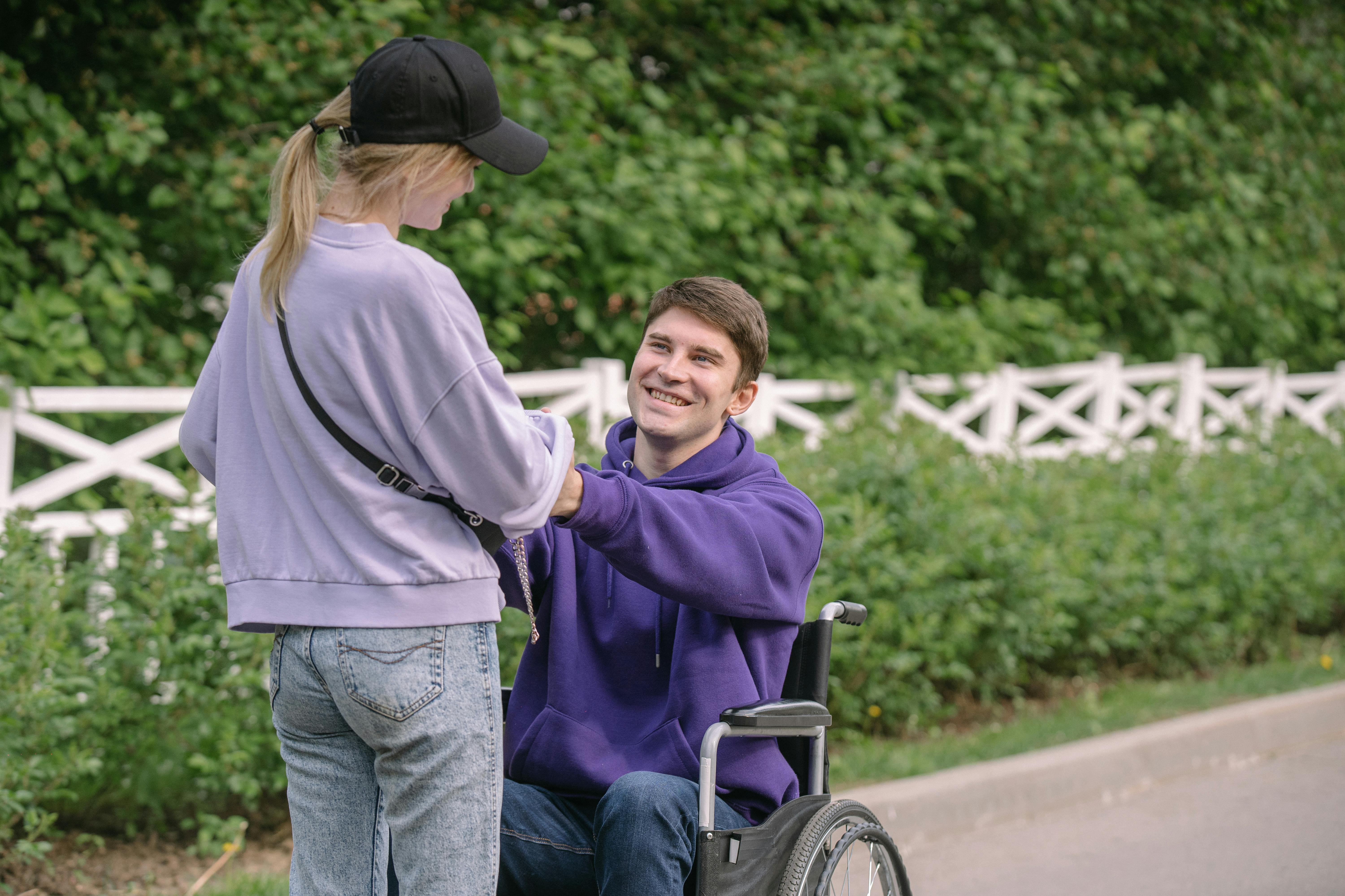A smiling young man in a wheelchair holding hands with a standing woman outdoors in a park. They appear to be enjoying each other's company, with greenery and a white fence in the background.