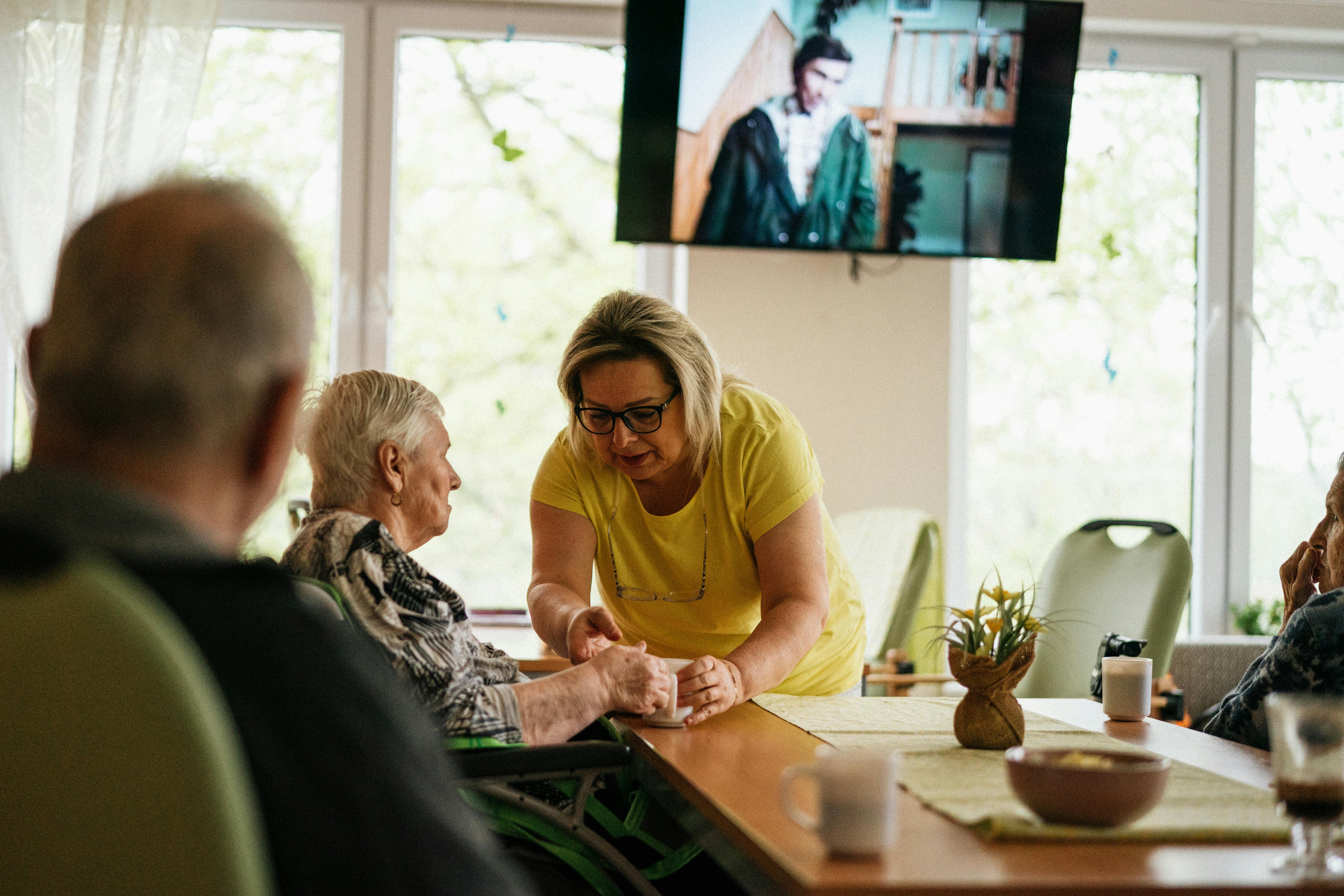 A blonde woman in a yellow shirt leans over a table, offering a cup to an elderly woman in a wheelchair, while other seniors are seated at the table in what appears to be a communal living or care setting, with a television screen displaying a person in the background.