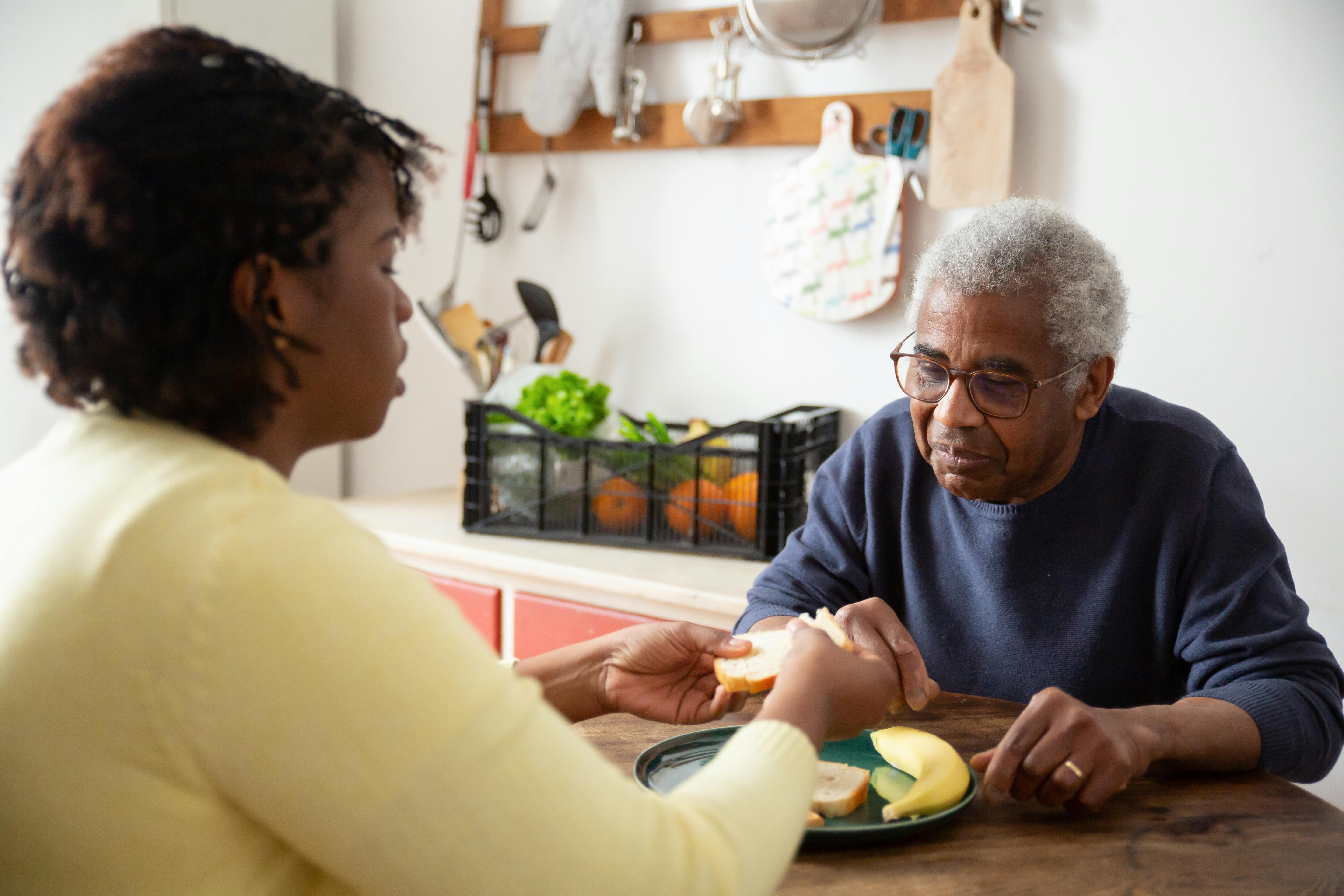 A woman in a yellow sweater assists an elderly man in a blue sweater with eating at a table, with fruit and bread on a plate between them.