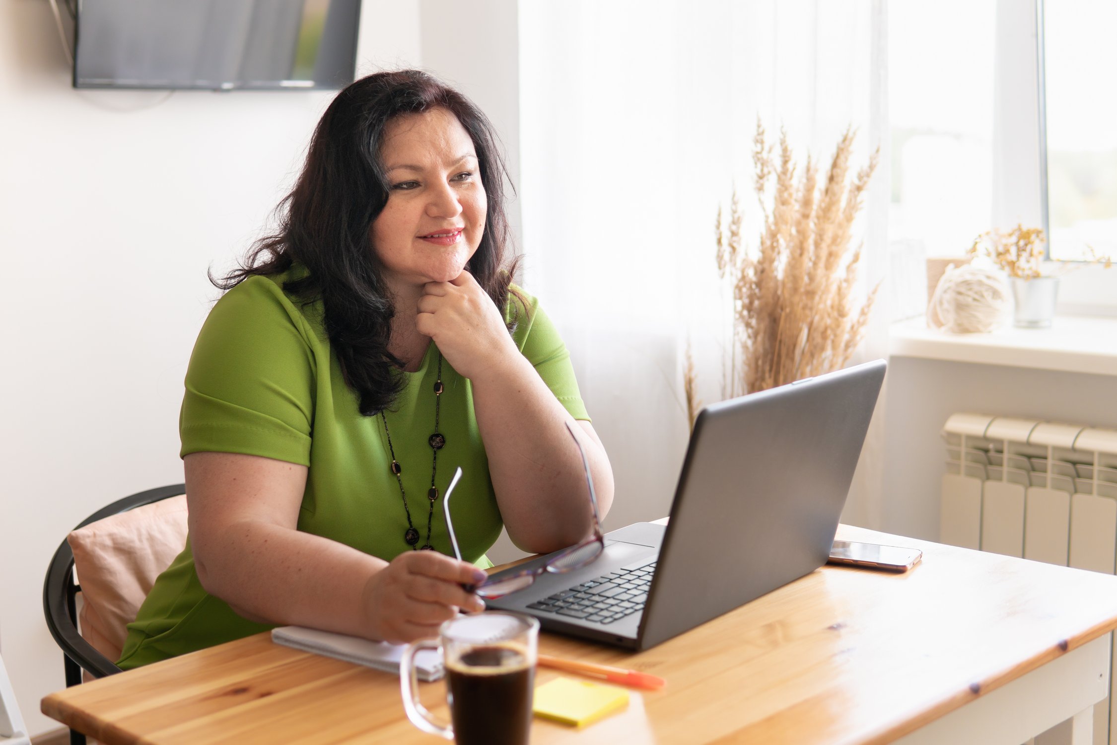 A middle-aged woman with dark hair and a green shirt, wearing glasses, sits at a wooden desk, looking at a laptop screen with a thoughtful expression, holding her chin. A cup of coffee and some office supplies are on the desk.