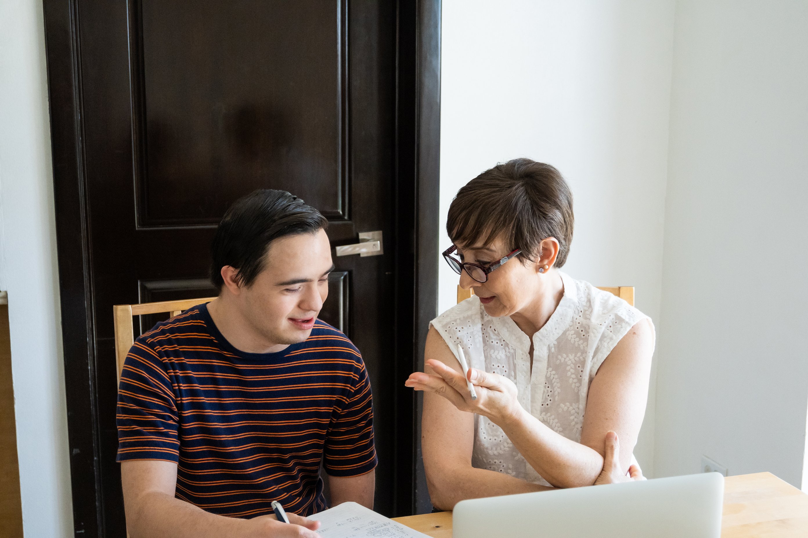 A woman with glasses and a man with learning disabilities are sitting at a table with a laptop and paper, appearing to be engaged in a learning or discussion session.