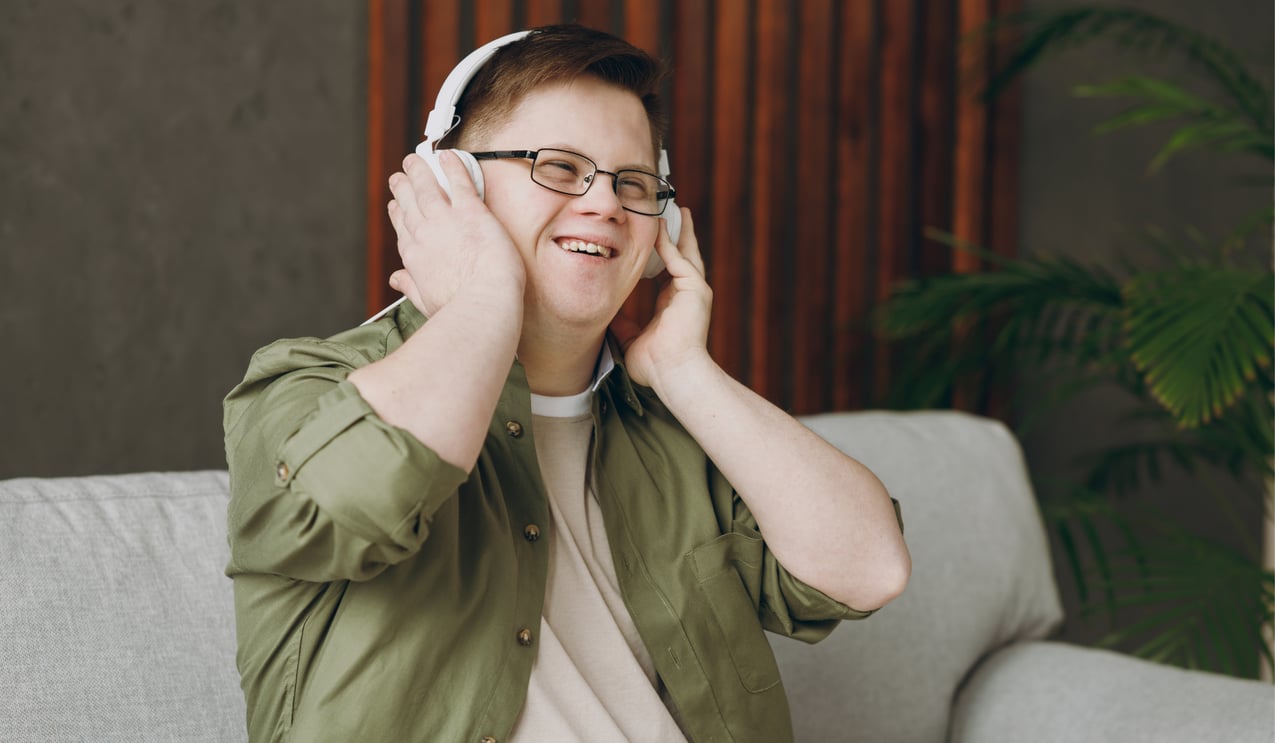 Young man with glasses and headphones, smiling on a gray couch with a plant in the background.