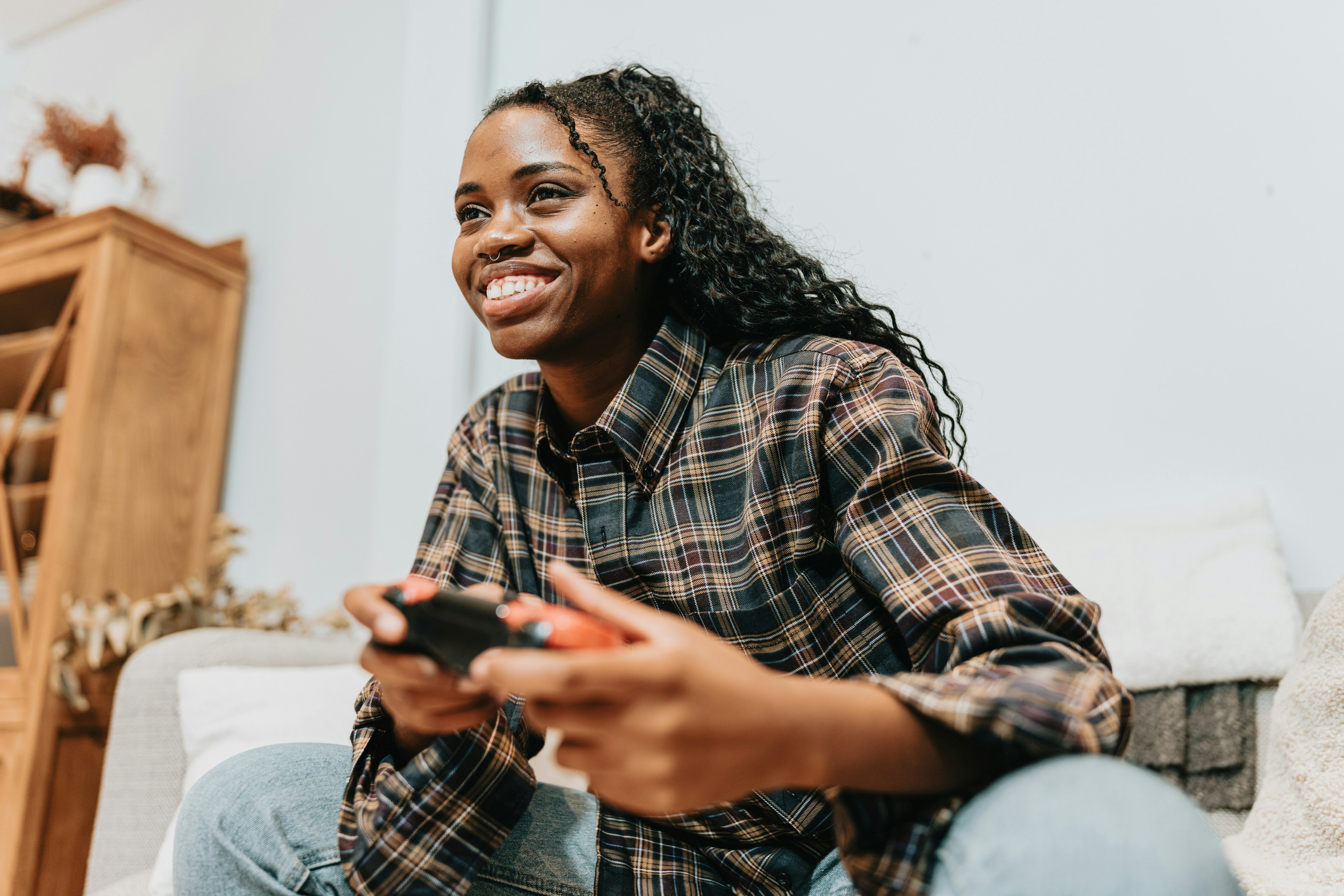 A young woman with long dark braids, wearing a plaid shirt and jeans, sitting on a couch and smiling while holding a video game controller. 