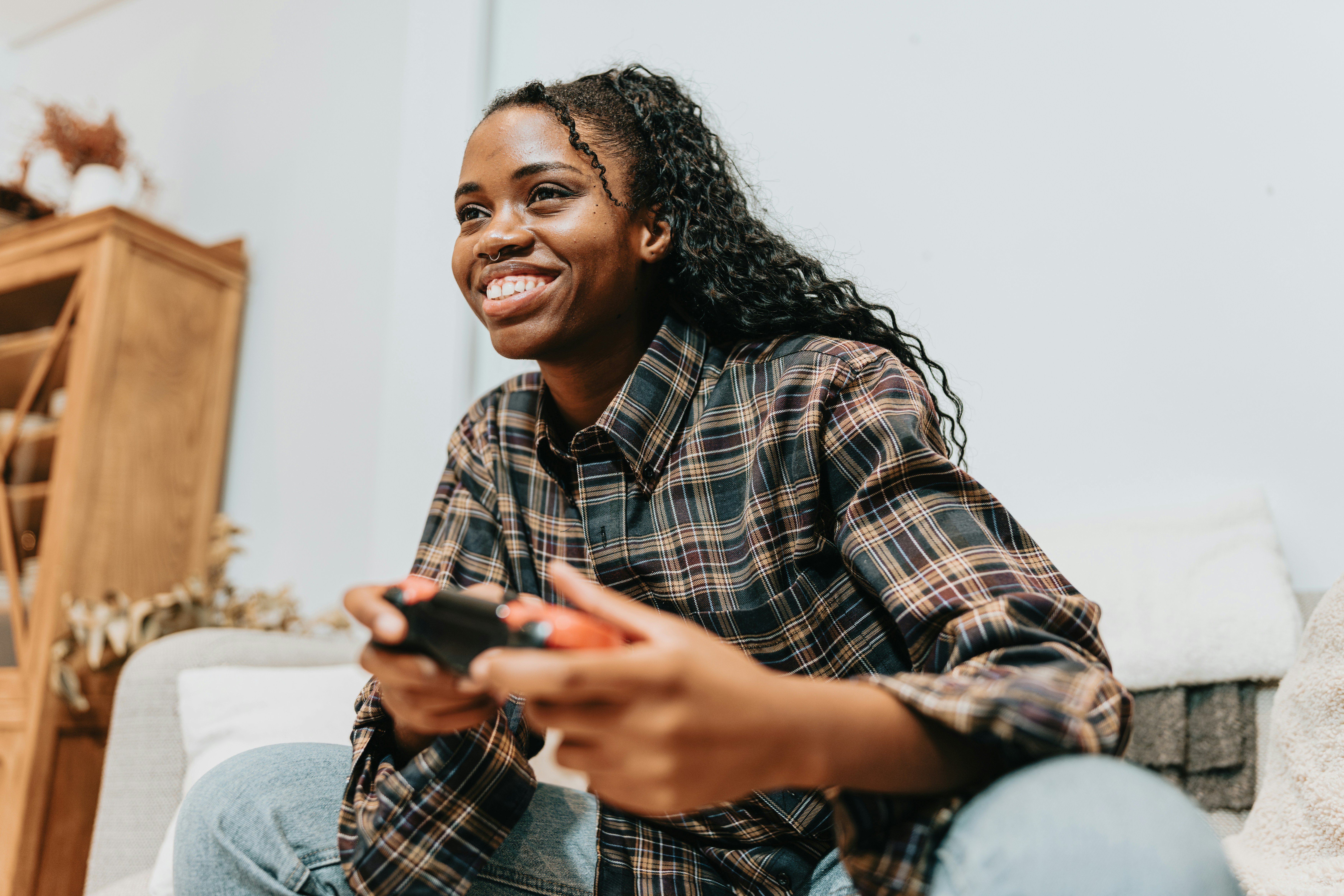A young woman with long dark braids, wearing a plaid shirt and jeans, sitting on a couch and smiling while holding a video game controller.