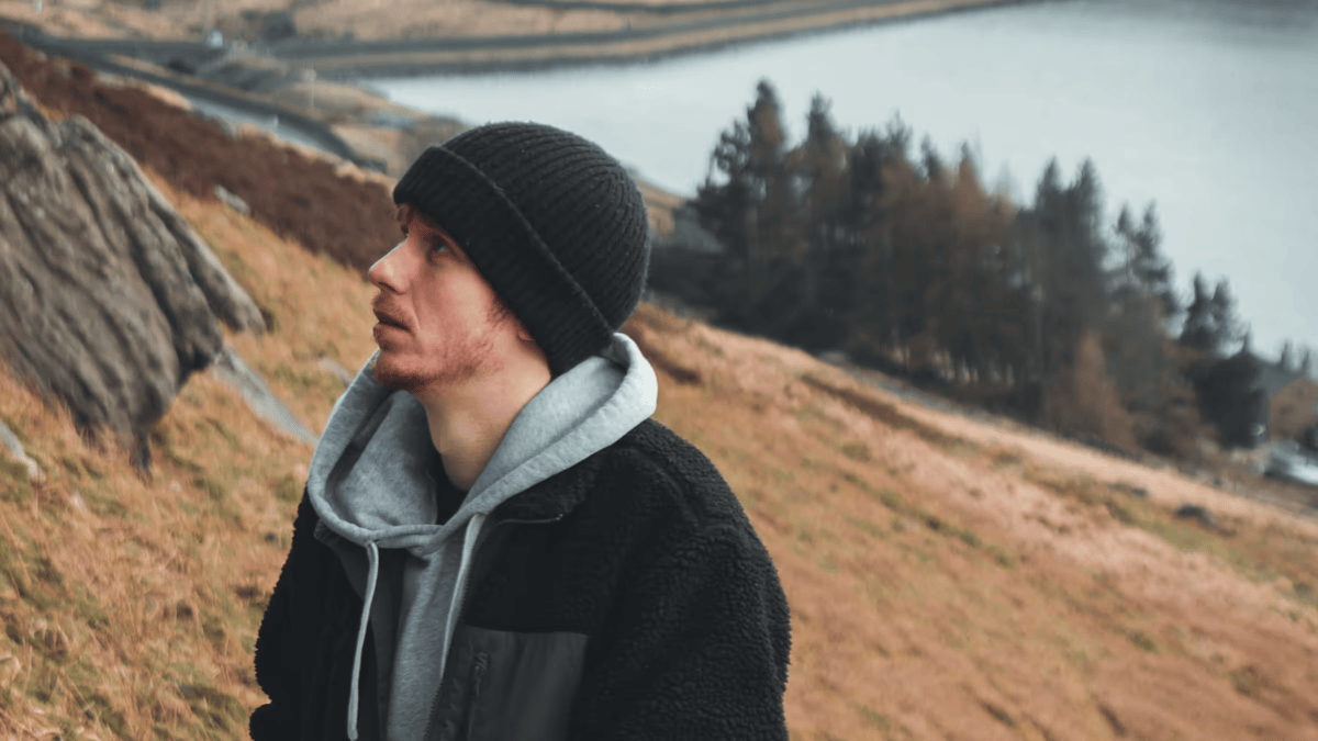 A young man in a black beanie, grey hoodie, and black jacket, with a reddish beard, looks up thoughtfully while standing on a grassy, hilly landscape overlooking a body of water and trees under an overcast sky.
