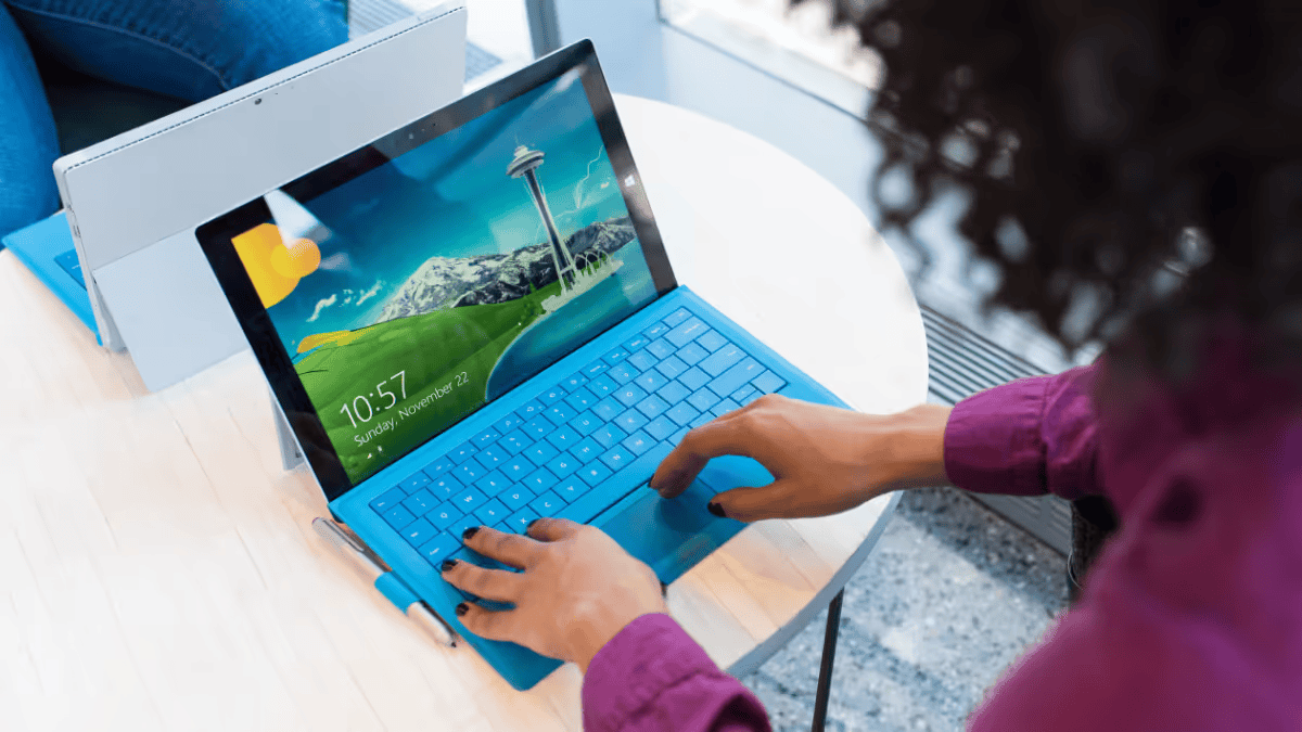 A person is using laptop with a bright blue keyboard. The laptop's screen shows the Windows lock screen. Another person is partially visible in the blurred background, seated at what appears to be another laptop.
