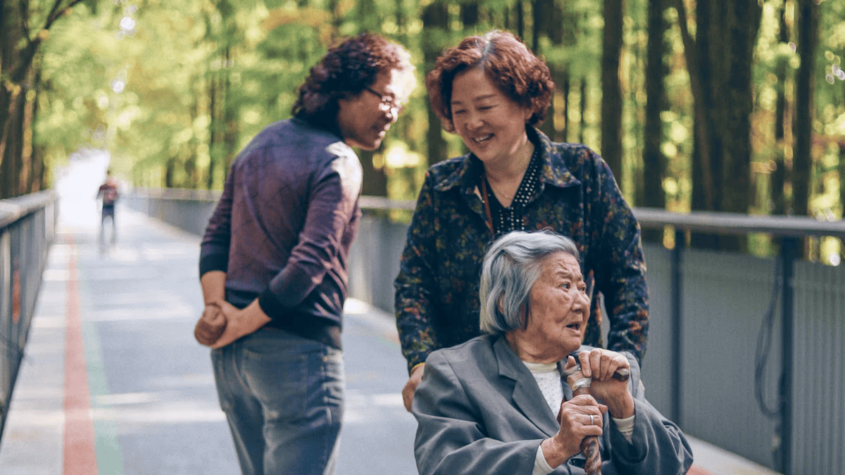 An elderly Asian woman in a wheelchair is smiling while looking up at two younger Asian individuals who are standing beside her and smiling back 