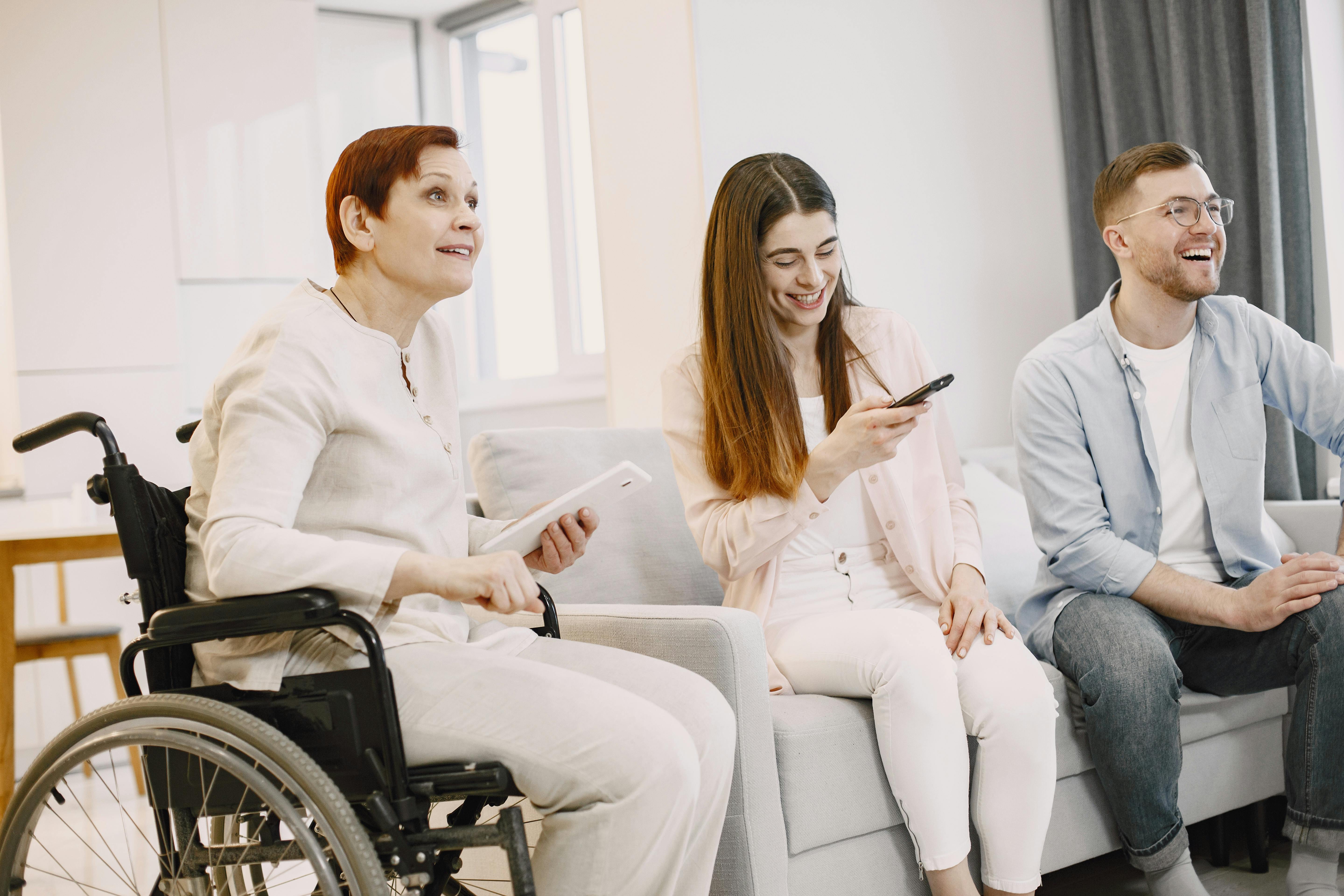 A woman in a wheelchair and a young couple are sitting in a living room, looking towards the right and smiling.