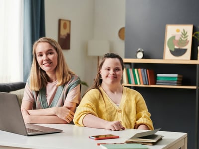 two people sitting at a table with a laptop, phone and notepad, one of them has downs syndrome